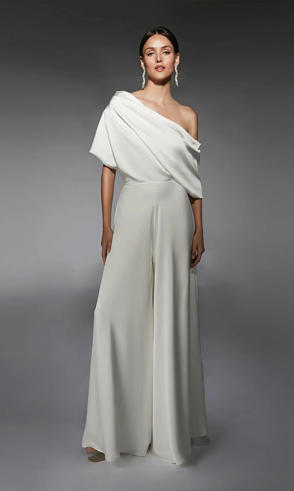 A woman poses in the Alyce 70072 long white jumpsuit in ivory with wide-leg pants and statement earrings, set against a plain gray background.
